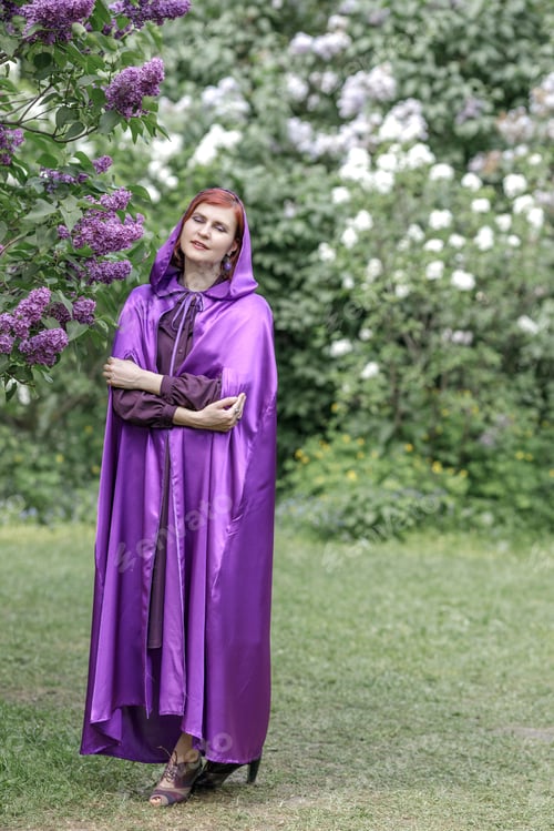 Preview: A beautiful young woman with red hair, against a background of blooming lilacs, in a cloak and hood