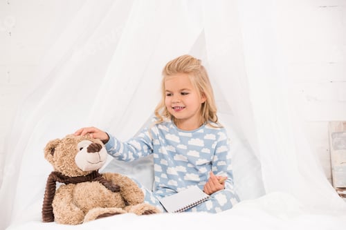 Preview: cute kid in pajamas sitting on bed with notebook and teddy bear