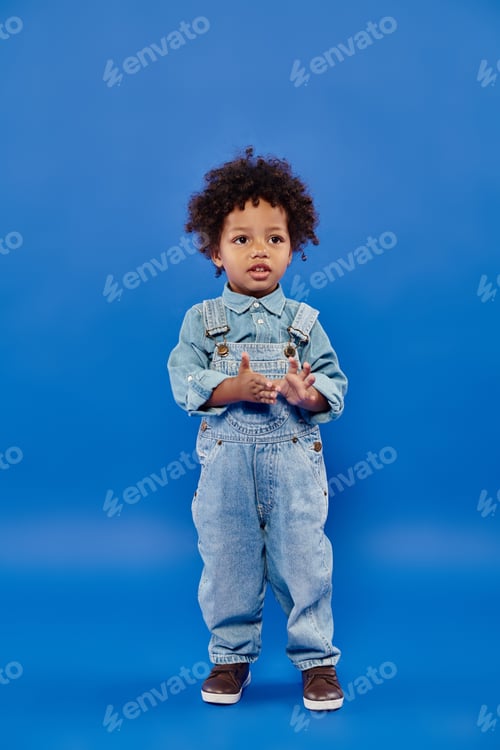 Preview: adorable and curly african american toddler boy in denim clothes standing on blue background
