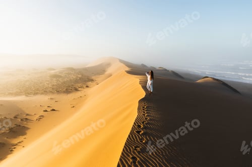 Preview: Woman enjoying sunrise on top of huge sand dune. Beautiful warm sun light and mist in morning Sahara