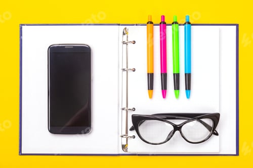 Preview: Top view of working space table with notebook, glasses and colorful pens isolated on yellow
