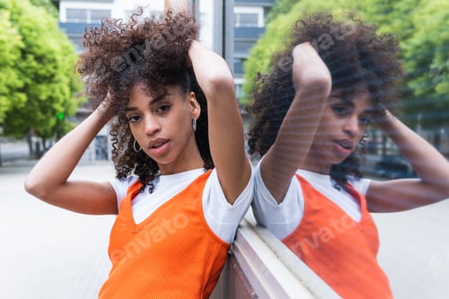 Preview: Ethnic woman touching curly hair against glass wall in city