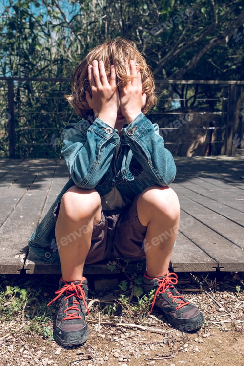 Preview: Upset boy sitting on wooden boardwalk and covering face