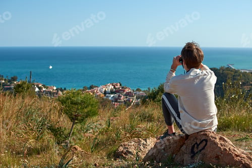 Preview: Back view of a young male sitting on a rock using binoculars looking at the sea