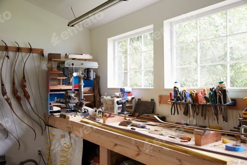 Preview: A workshop bench with components and tools, and finished articulated bows hanging on the wall.