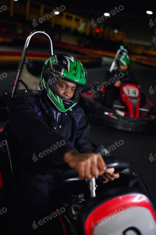 Preview: african american driver in helmet driving go kart on indoor circuit near friend on blurred backdrop