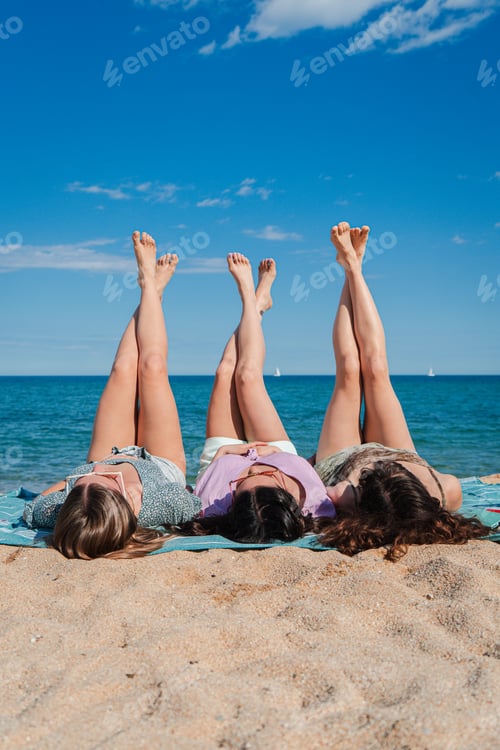 Preview: Vertical Group of feminine girls lifting their legs lying on a beach towel in the sand. Three young