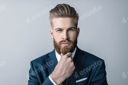 Preview: Stylish bearded businessman with hand on chin looking at camera isolated on grey