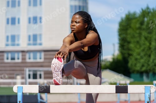Preview: Black young woman with long braids stretches legs and body using hurdles barrier on sports ground