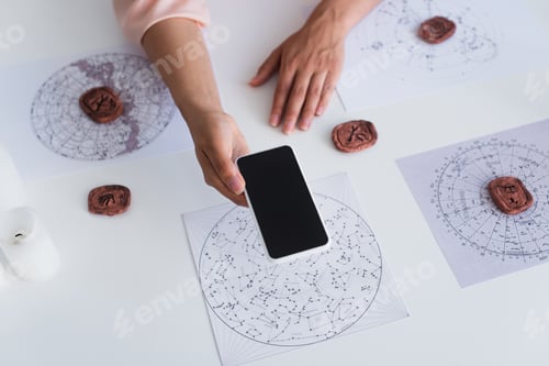 Preview: top view of cropped astrologist holding mobile phone with blank screen near celestial charts and