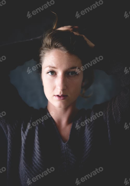 Preview: Woman with intense gaze tying her hair up in a dimly lit room.