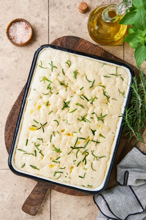 Preview: Raw focaccia dough inside baking dish with ingredients, olive oil and basil on rustic old table. Pro