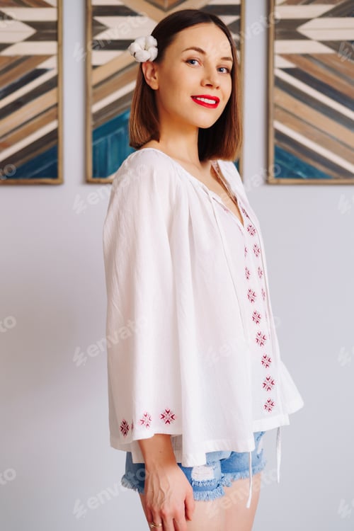 Preview: Smiling woman portrait with red lips, wearing folk blouse and cotton flower in her hair indoors