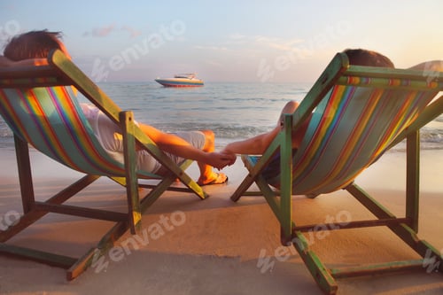 Preview: Happy couple sitting at sun chairs on the beach of Koh Samet at sunset