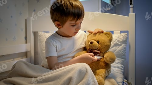 Preview: Portrait of cute sick boy measuring temperature to his toy teddy bear with digital thermometer