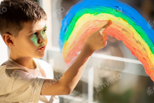 Preview: Serious little boy draws rainbow on window with hand, dream, think in room interior, sun flare