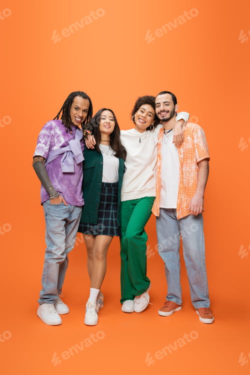 Preview: full length of happy african american woman embracing stylish multicultural friends on orange