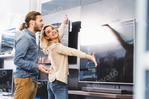 Preview: smiling girlfriend pointing with hands at tv and looking at shocked boyfriend in home appliance
