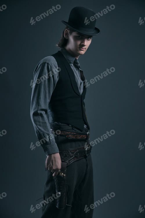 Preview: Western 1900 Fashion Man With Brown Hair And Hat Holding Gun. Studio Shot Against Grey.