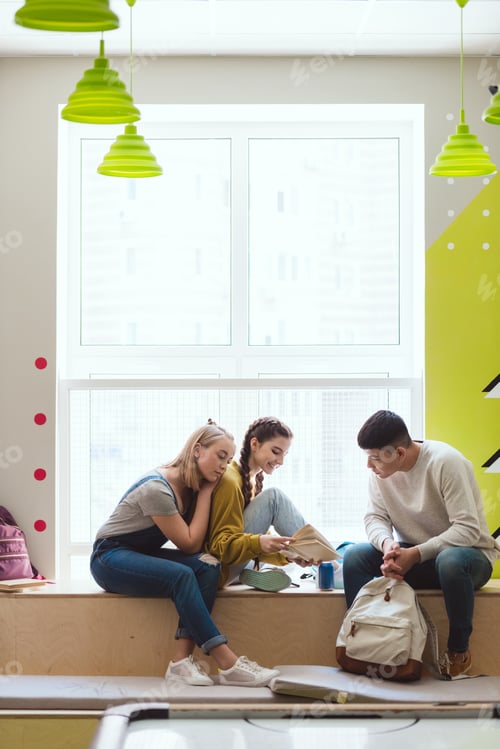 Preview: Multicultural group of teenage schoolchildren sitting on window sill and reading book