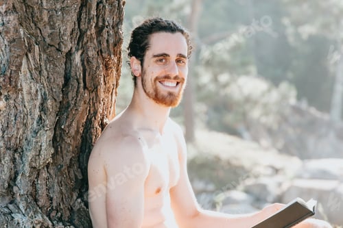 Preview: Close up of a man with a beard holding a book and smiling to camera shirtless next to a tree