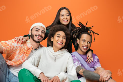 Preview: young asian woman embracing cheerful multicultural friends looking away isolated on orange