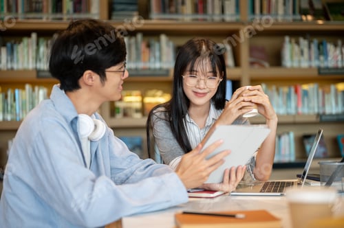 Preview: Glasses asian man student holding tablet showing to woman friend sits with laptop on library's table