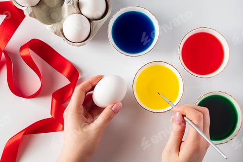 Preview: Young woman paints white eggs for Easter on white background. The egg in hand, flat lay