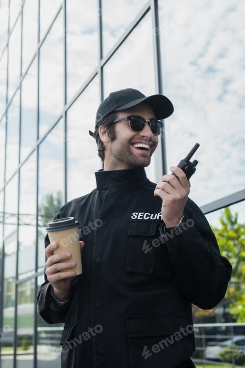 Preview: cheerful security man with paper cup talking on walkie-talkie on city street