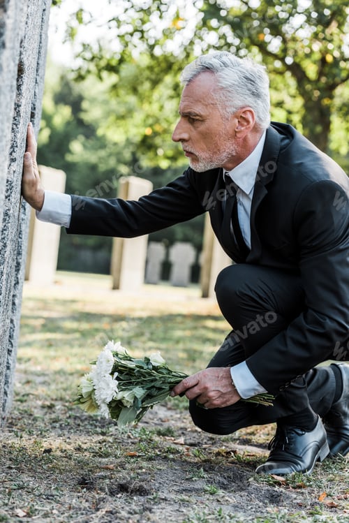 Preview: upset man with grey hair putting flowers near tomb in cemetery