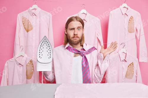 Preview: Puzzled bearded redhead man uses iron to get wrinkles out of shirt raises palm while standing near i