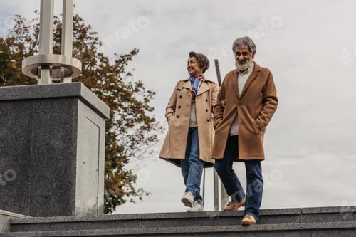 Preview: full length of stylish senior man in coat walking on stairs with hands in pockets near happy wife