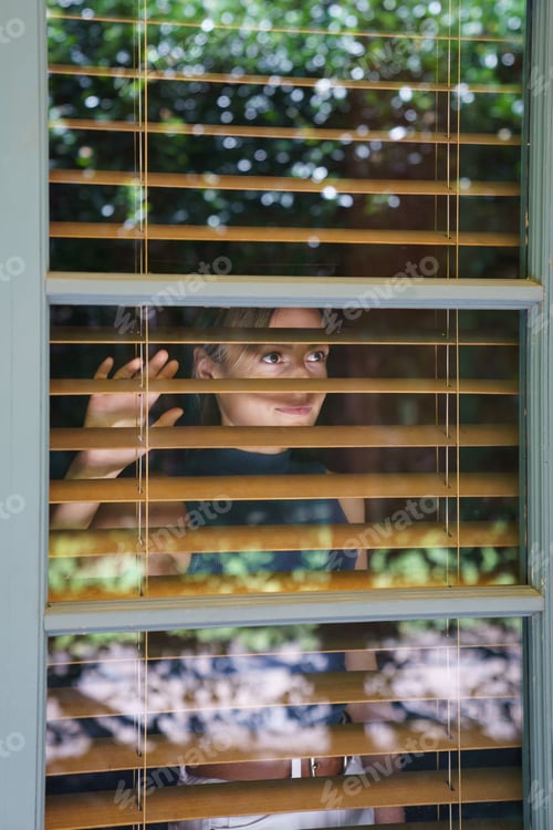 Preview: Young woman enjoys summer nature on sunny day looking through brown shutter on big window of house