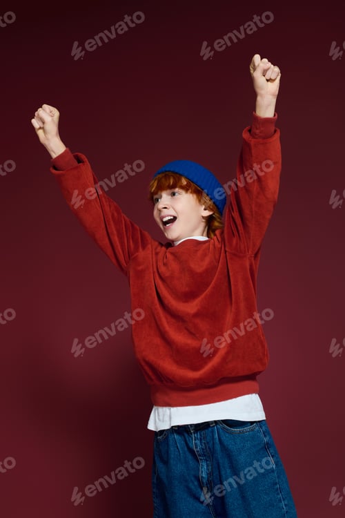Preview: Joyful redhead boy celebrates in stylish casual outfit in a vibrant studio setting