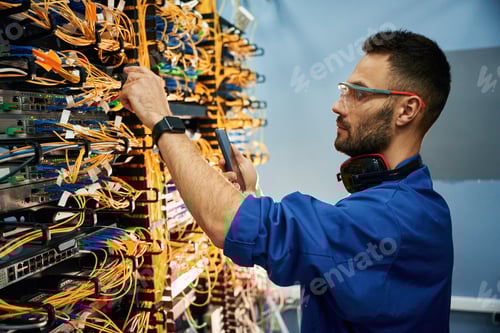 Preview: Side view. Young man is working with internet equipment and wires in server room