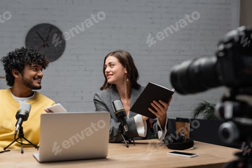 Preview: cheerful brunette podcaster in grey blazer sitting with notebook near young indian colleague