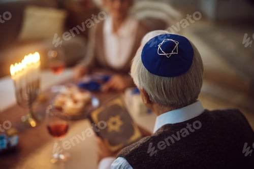 Preview: Close up of senior Jewish man reading Tanakh while celebrating Hanukkah with his wife at home.