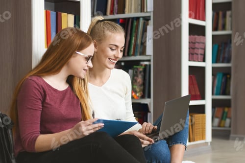 Preview: Cheerful teenage girlfriends sitting on floor in library, using laptop