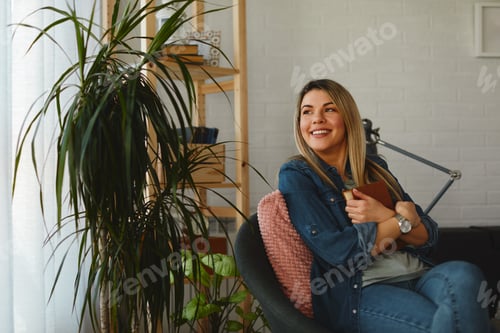 Preview: Young beautiful woman sitting in a chair by the window and holding a book in her hands