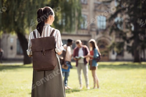Preview: back view of young female student with backpack looking at classmates standing on grass near