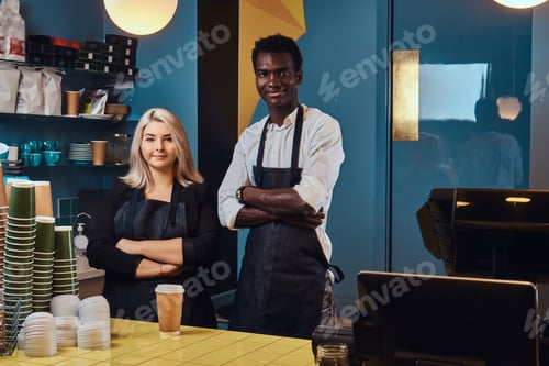 Preview: Two young multiracial baristas in aprons standing welcomingly at their trendy coffee shop.