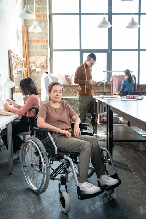 Preview: Young disable seamstress in casualwear sitting in wheelchair in front of camera against colleagues