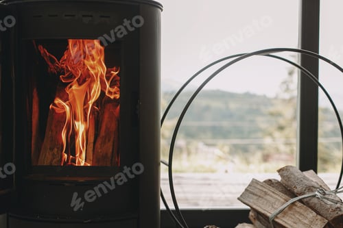 Preview: Modern black fireplace with fire and firewood on metal stand at window with view on mountains
