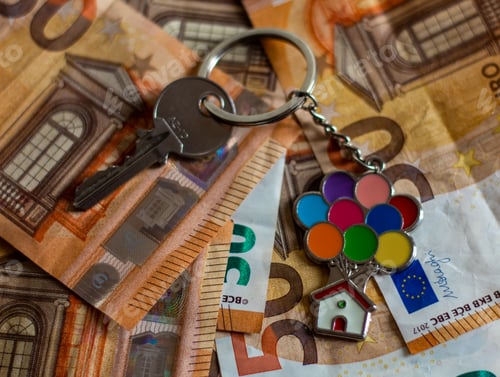 Preview: Metal key with a keychain of a house with colorful balloons, on top of a pile of euro bills.