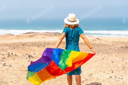 Preview: A lesbian person with a green dress, a white hat and with the LGBT flag by the sea