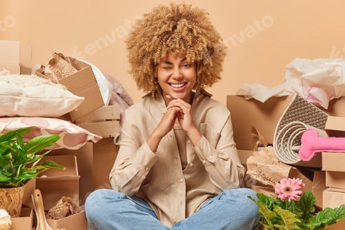 Preview: Moving day. Positive woman with curly bushy hair keeps hands under chin winks eye sits on floor