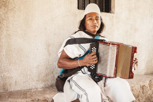 Preview: An indigenous Arhuaco man in traditional dress plays the accordion seated on a stone bench.