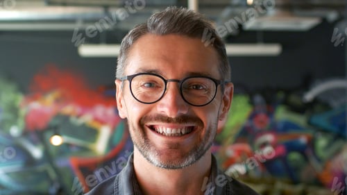 Preview: Portrait Of Smiling Mature Man Wearing Glasses In Urban Setting With Graffiti Covered Wall Behind