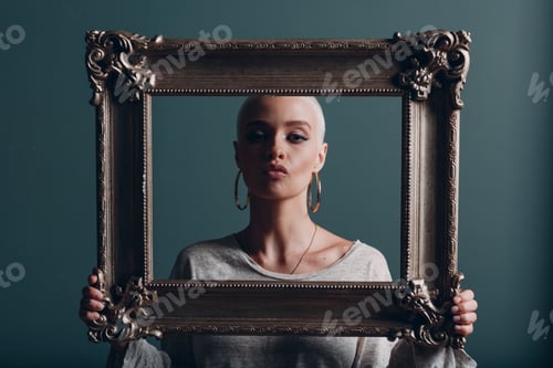 Preview: Millenial young woman with short blonde hair holds gilded picture frame in hands behind her face por