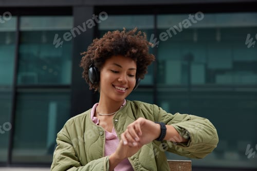 Preview: Horizontal shot of good looking Afro American woman with curly hair checks time on smartwatch listen